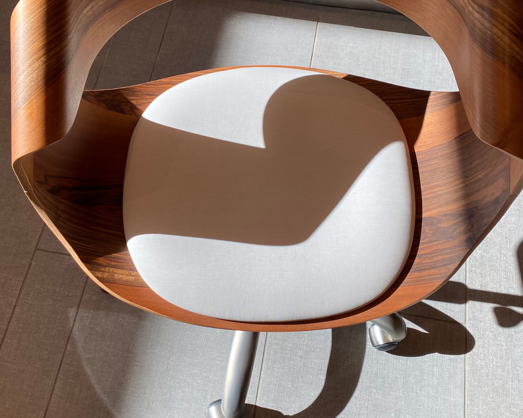 Sunlit ergonomic chair with curved wooden backrest and white cushioned seat, casting a soft shadow on a tiled floor.