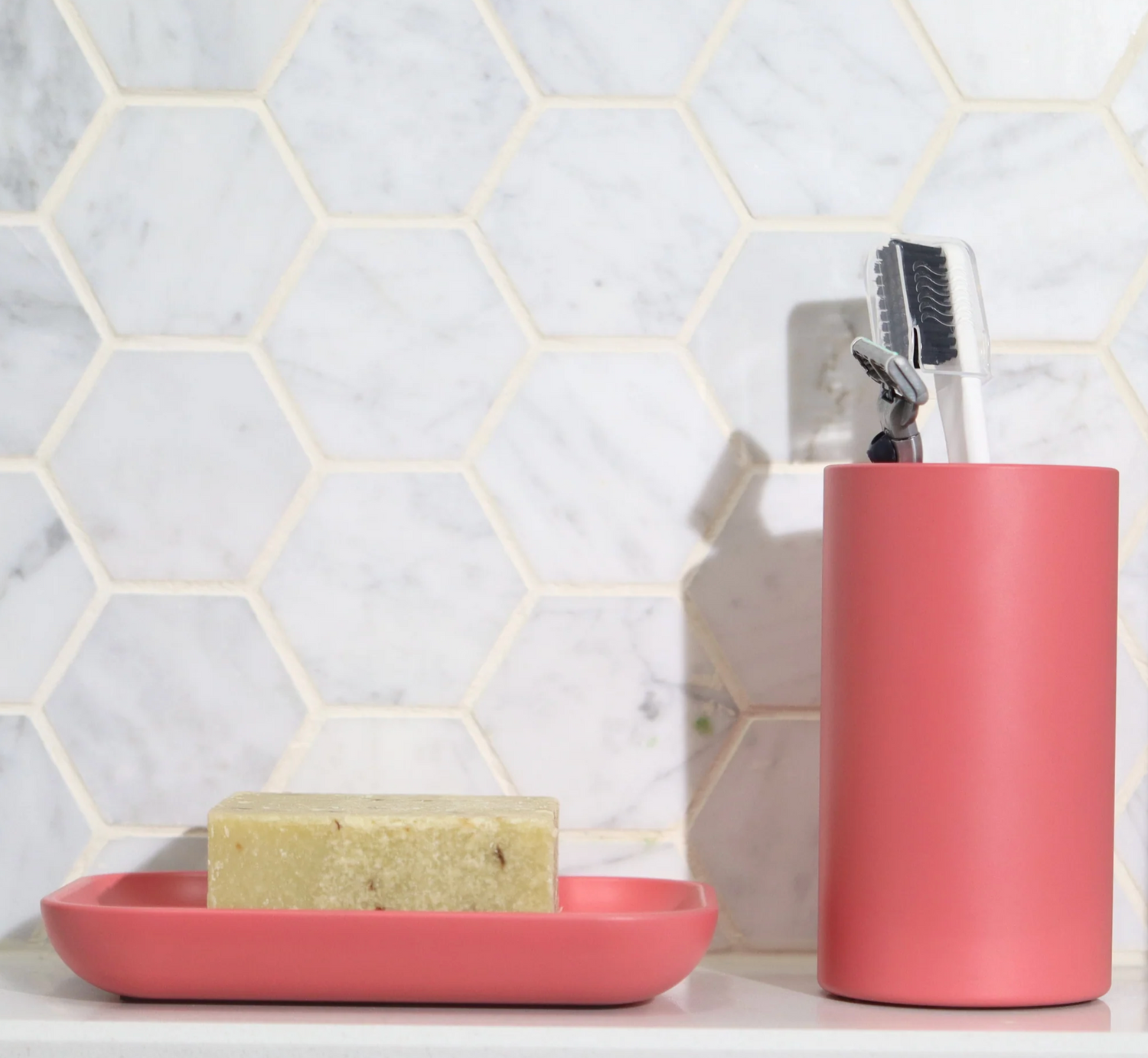 Pink soap dish holding a bar of soap and a matching toothbrush holder with a toothbrush and razor, set against a tile wall.